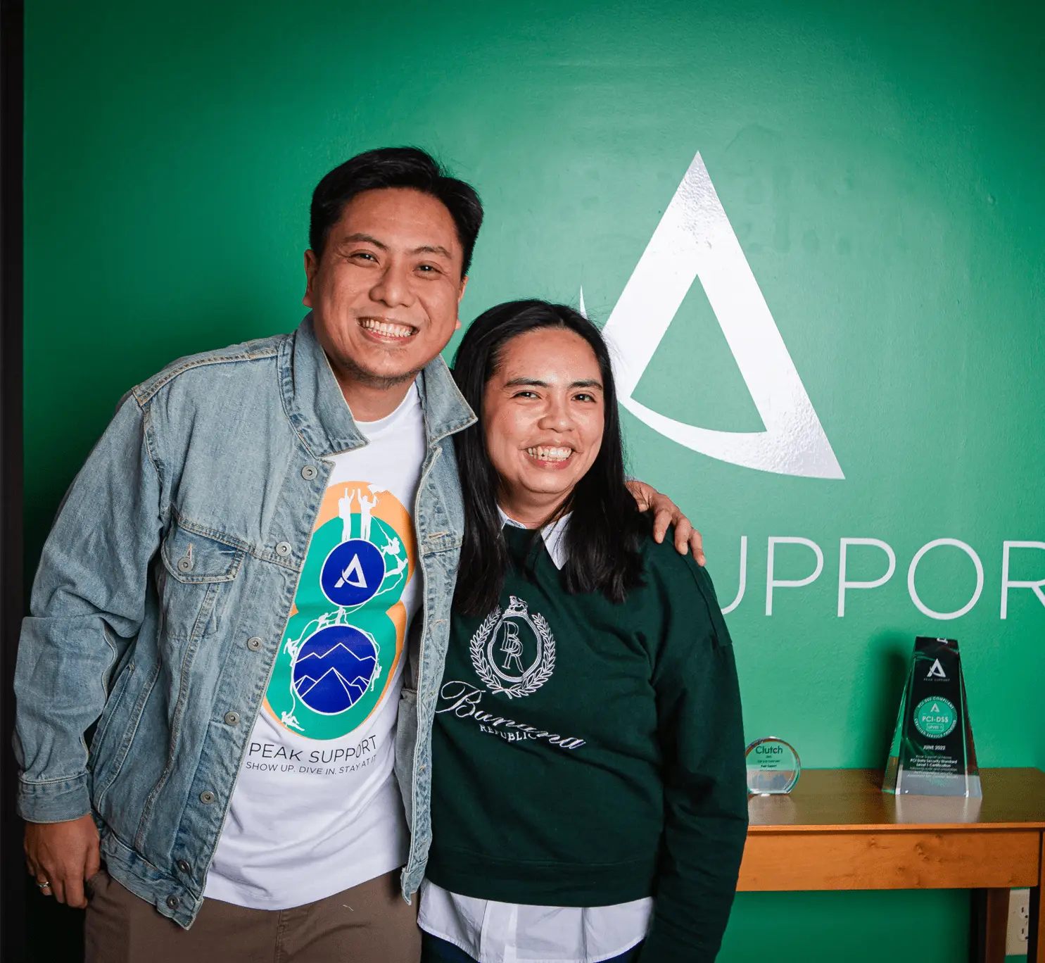 Two Peak Support team members smile in front of the company logo, representing the people-first culture that powers exceptional customer service. Peak Support helps companies scale with people-first CX solutions.